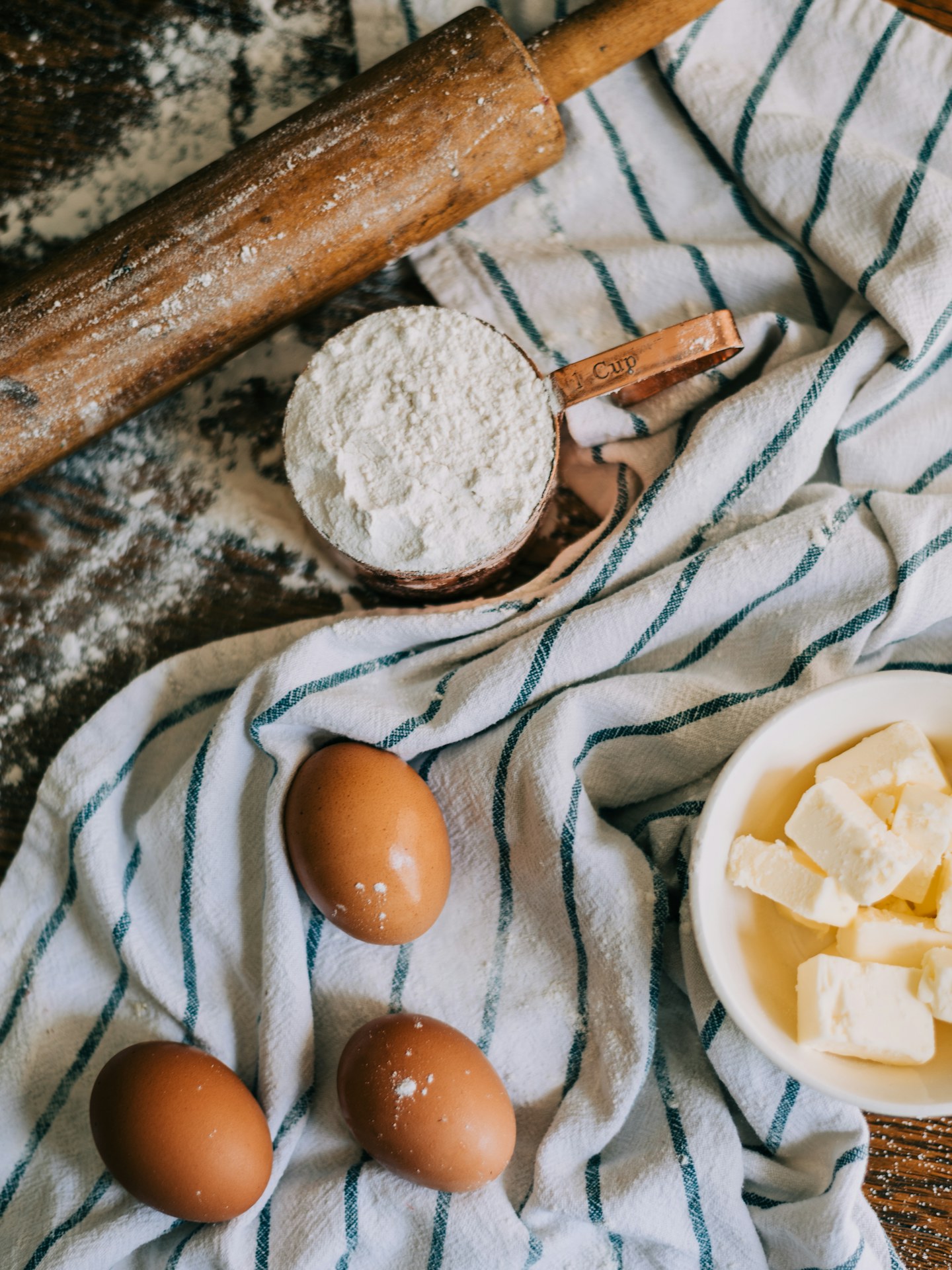 Flour, eggs, butter, and a rolling pin arranged on a striped cloth.
