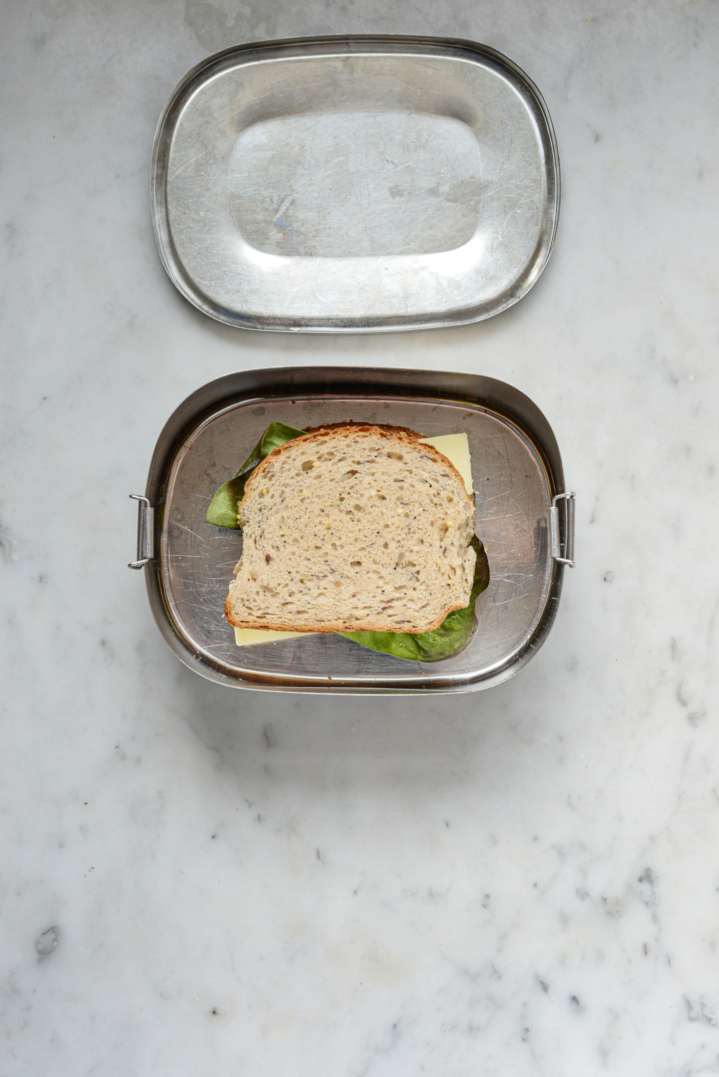 A sandwich packed in an open metal lunch container on a stone surface.